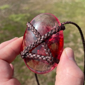 May include: A red, clear, oval-shaped pendant with a brown leather cord wrapped around it. The pendant is translucent and has a slight shimmer.