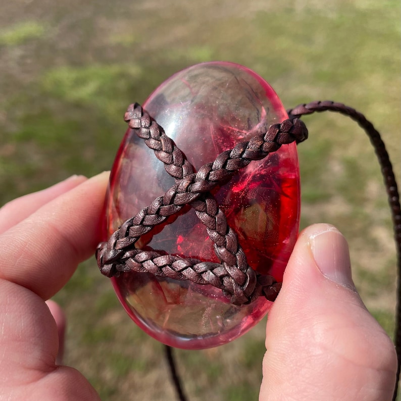 May include: A red, clear, oval-shaped pendant with a brown leather cord wrapped around it. The pendant is translucent and has a slight shimmer.