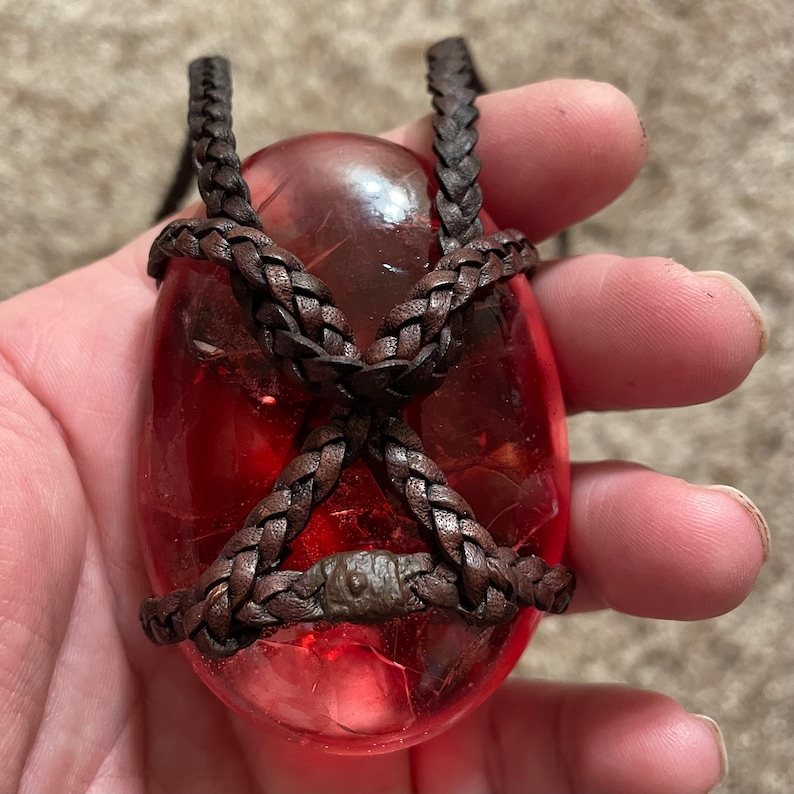 May include: A red glass pendant with a brown leather cord wrapped around it. The cord is braided and has a knot at the top.