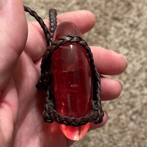 May include: A red glass pendant wrapped in brown leather cord. The pendant is shaped like a teardrop and has a smooth, polished surface.