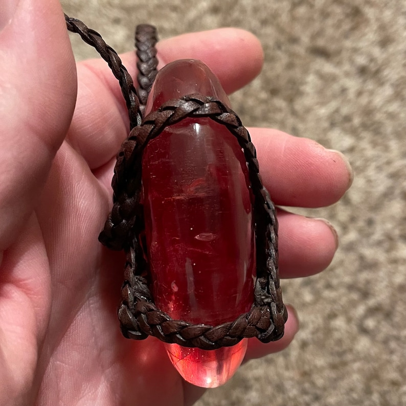 May include: A red glass pendant wrapped in brown leather cord. The pendant is shaped like a teardrop and has a smooth, polished surface.