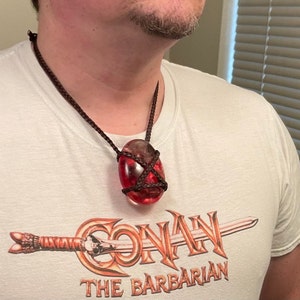 May include: A red, oval-shaped pendant with a brown leather cord. The pendant is wrapped with a brown leather cord and has a clear, glassy appearance. The pendant is hanging from a brown leather cord around a person's neck. The person is wearing a white t-shirt with the words "Conan The Barbarian" printed on it.
