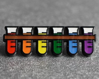 May include: A set of seven enamel pins shaped like test tubes, each a different color of the rainbow. The pins are arranged in a row on a brown surface.