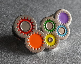 May include: A silver pin with six interlocking gears, each with a different colored center: red, orange, yellow, green, blue, and purple.
