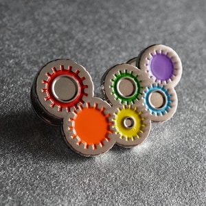 May include: A silver pin featuring six interlocking gears, each with a different coloured centre: red, orange, yellow, green, blue, and purple.