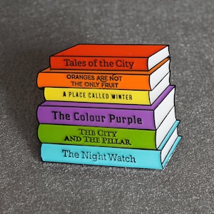 Könnte beinhalten: Ein Stapel Emaille-Pins, die Buchcover darstellen. Die Pins sind in Regenbogenfarben gehalten und zeigen die Titel "Tales of the City", "Oranges Are Not the Only Fruit", "A Place Called Winter", "The Colour Purple", "The City and the Pillar" und "The Night Watch".