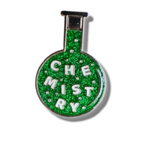 Può includere: Uno spillo in smalto verde glitterato a forma di becher con la parola "CHIMICA" scritta in lettere bianche.