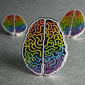 Könnte beinhalten: Drei Gehirn-Emaille-Anstecknadeln. Die Anstecknadeln haben einen schwarzen Umriss mit einem Regenbogenfarbenen Gehirndesign. Das Gehirn ist in zwei Hälften geteilt, jede mit einem anderen Farbschema. Die Anstecknadeln sind vor einem grauen Hintergrund platziert.