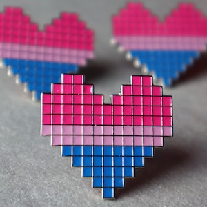May include: Three pixelated heart-shaped pins in pink, light pink, and blue colors. The pins are arranged in a triangular pattern on a gray surface.