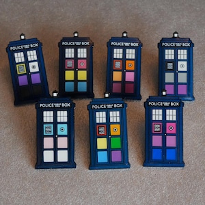 May include: Seven blue Police Box magnets, each with a different arrangement of coloured panels. The magnets display the text "POLICE PUBLIC CALL BOX" and are arranged on a neutral surface, reminiscent of the iconic British phone box.