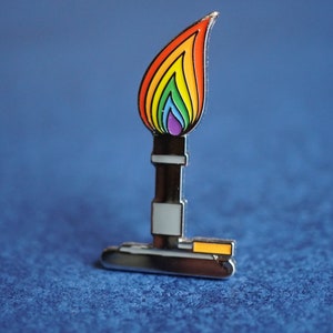 May include: A rainbow-coloured flame pin with a silver base and a yellow accent. The pin is a representation of a Bunsen burner.