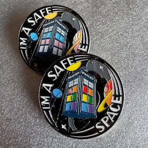May include: Two black enamel pins featuring a Tardis design with rainbow-coloured windows. The pins have the text "I'M A SAFE SPACE" in white. The design includes planets, stars, and a yellow sun. The Tardis is a blue police box.