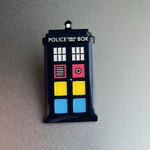 Pode incluir: Um &iacute;man azul marinho da cabine da pol&iacute;cia com as palavras "POLICE PUBLIC CALL BOX" no topo. O &iacute;man tem janelas brancas, um quadrado vermelho com um c&iacute;rculo branco e quadrados amarelos e azuis.