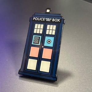Pode incluir: Um alfinete de esmalte azul em forma de cabine telef&oacute;nica da pol&iacute;cia. O alfinete apresenta as palavras "POLICE PUBLIC BOX" no topo. A porta tem pain&eacute;is brancos, rosa e azuis. O alfinete &eacute; um item de colec&ccedil;&atilde;o.