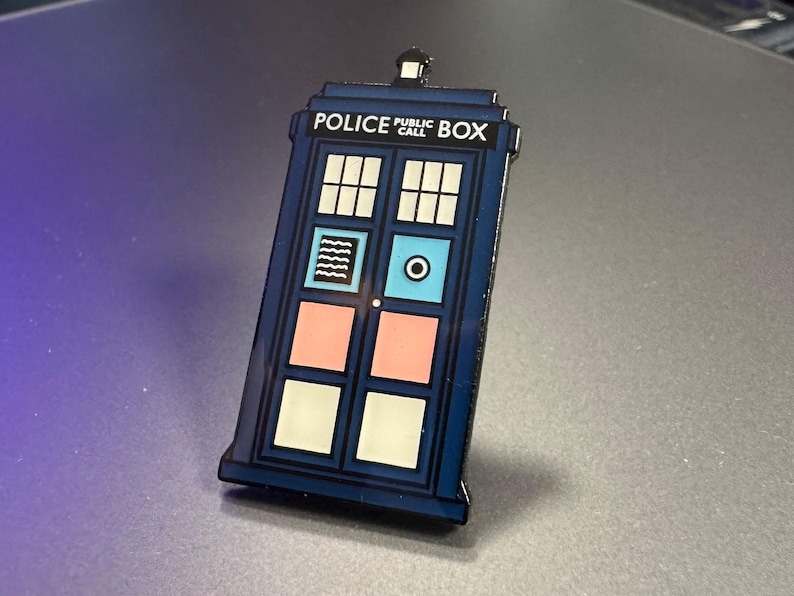 Pode incluir: Um alfinete de esmalte azul em forma de cabine telef&oacute;nica da pol&iacute;cia. O alfinete apresenta as palavras "POLICE PUBLIC BOX" no topo. A porta tem pain&eacute;is brancos, rosa e azuis. O alfinete &eacute; um item de colec&ccedil;&atilde;o.