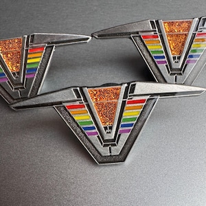 Puede incluir: Tres pines plateados con un diseño en forma de V. Los pines presentan un patrón de rayas de arcoíris y un centro naranja brillante. Los pines tienen un estilo geométrico art déco.