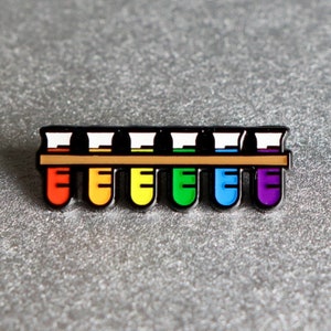K&ouml;nnte beinhalten: Eine Regenbogen-Emaille-Anstecknadel mit einer Reihe von acht Reagenzgl&auml;sern, die jeweils mit einer anderen Farbe Fl&uuml;ssigkeit gef&uuml;llt sind. Die Reagenzgl&auml;ser sind in einem Regenbogenmuster angeordnet, mit Rot, Orange, Gelb, Gr&uuml;n, Blau, Indigo und Violett.