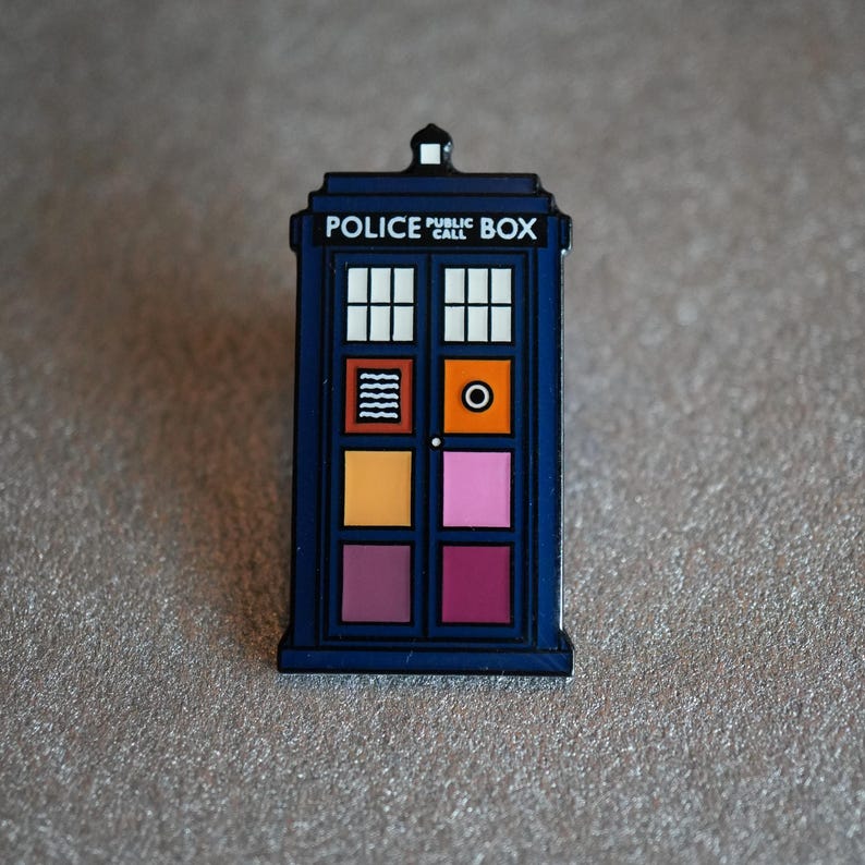 Pode incluir: Um alfinete azul da caixa telef&oacute;nica da pol&iacute;cia com formato retangular. O alfinete apresenta um design de janela branca e uma s&eacute;rie de quadrados coloridos em laranja, rosa e roxo. O texto "POLICE PUBLIC CALL BOX" est&aacute; impresso no topo.