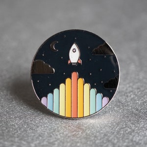Peut inclure: Épingle ronde en émail représentant une fusée s'élevant dans un ciel étoilé. L'épingle présente une traînée de lancement arc-en-ciel et un croissant de lune. La broche mesure environ 3,2 cm de diamètre.