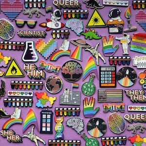K&ouml;nnte beinhalten: Eine Sammlung von Emaille-Pins mit verschiedenen Designs, darunter Regenb&ouml;gen, Planeten, Dinosaurier und wissenschaftliche Motive. Die Pins sind auf einem violetten Hintergrund angeordnet und zeigen die W&ouml;rter "Queer", "Scientist", "He Him", "They Them", "She Her" und "Doc".