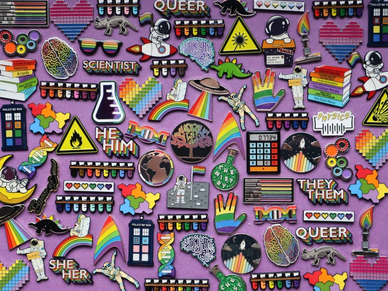 K&ouml;nnte beinhalten: Eine Sammlung von Emaille-Pins mit verschiedenen Designs, darunter Regenb&ouml;gen, Planeten, Dinosaurier und wissenschaftliche Motive. Die Pins sind auf einem violetten Hintergrund angeordnet und zeigen die W&ouml;rter "Queer", "Scientist", "He Him", "They Them", "She Her" und "Doc".