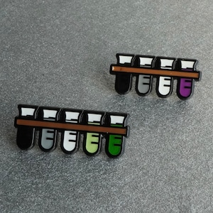 Op de afbeelding: Twee emaille pins met een rij van vijf reageerbuizen in een houten rek. De reageerbuizen zijn zwart, grijs, wit, groen en paars. De pins liggen op een getextureerd grijs oppervlak.