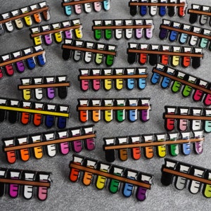 K&ouml;nnte beinhalten: Eine Sammlung bunter Emaille-Pins mit Reihen von Reagenzgl&auml;sern. Die Pins sind in verschiedenen Mustern und Designs angeordnet, wobei jede Reihe eine andere Farbkombination aufweist. Die Pins sind perfekt, um jedem Outfit einen Hauch von Verspieltheit und Pers&ouml;nlichkeit zu verleihen.