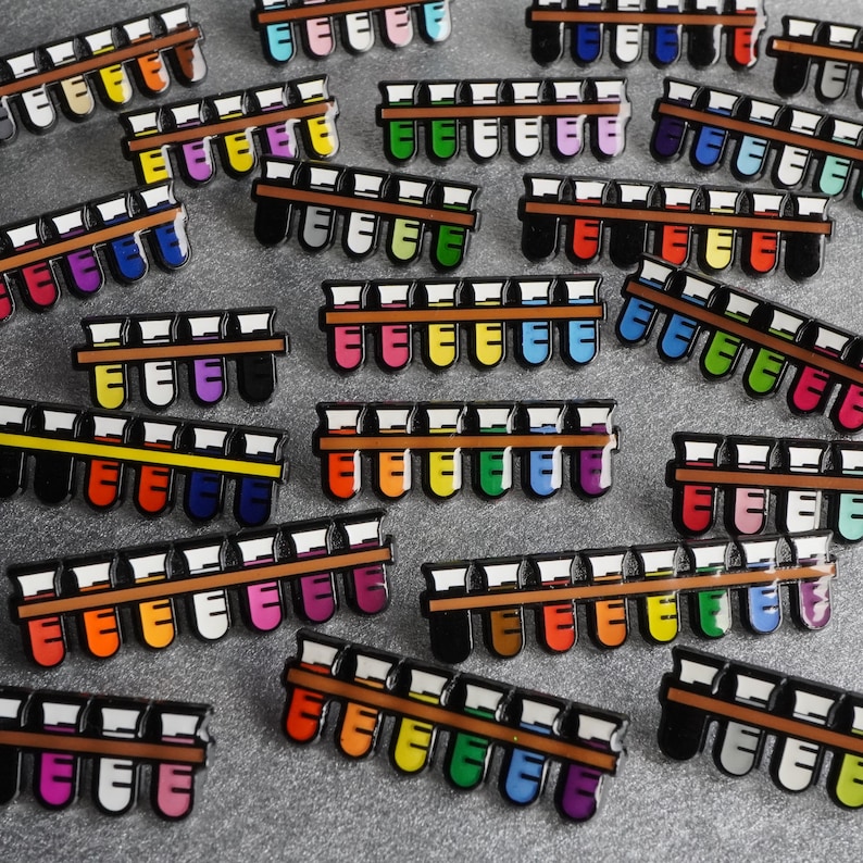 K&ouml;nnte beinhalten: Eine Sammlung bunter Emaille-Pins mit Reihen von Reagenzgl&auml;sern. Die Pins sind in verschiedenen Mustern und Designs angeordnet, wobei jede Reihe eine andere Farbkombination aufweist. Die Pins sind perfekt, um jedem Outfit einen Hauch von Verspieltheit und Pers&ouml;nlichkeit zu verleihen.