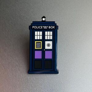 Pode incluir: Um &iacute;man azul de cabine telef&oacute;nica da pol&iacute;cia com as palavras "POLICE PUBLIC CALL BOX" no topo. O &iacute;man tem vidros brancos, um design amarelo e preto, um c&iacute;rculo branco e pain&eacute;is roxos e pretos nas portas.
