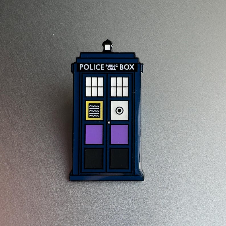 Pode incluir: Um &iacute;man azul de cabine telef&oacute;nica da pol&iacute;cia com as palavras "POLICE PUBLIC CALL BOX" no topo. O &iacute;man tem vidros brancos, um design amarelo e preto, um c&iacute;rculo branco e pain&eacute;is roxos e pretos nas portas.