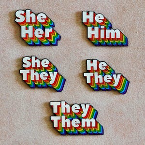 May include: Five enamel pins featuring white text with a rainbow-coloured drop shadow effect. The pins display the pronouns She/Her, He/Him, She/They, He/They, and They/Them. Each pin has a layered, multi-coloured border design.
