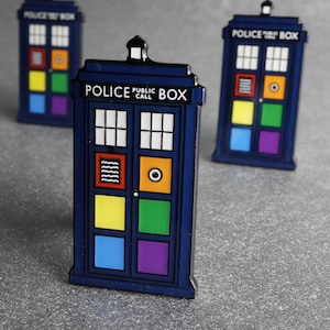 Pode incluir: Tr&ecirc;s pins azuis em forma de cabine telef&oacute;nica da pol&iacute;cia, com a inscri&ccedil;&atilde;o "POLICE PUBLIC CALL BOX". Cada pin apresenta pain&eacute;is coloridos com diferentes desenhos, incluindo um olho e linhas onduladas, sobre um fundo cinzento.