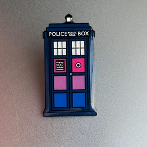 Pode incluir: Um &iacute;man azul da cabine telef&oacute;nica com o texto "POLICE PUBLIC CALL BOX". O &iacute;man apresenta um design retangular com janelas brancas, um c&iacute;rculo rosa e linhas onduladas. Os pain&eacute;is das portas s&atilde;o azuis e rosa.