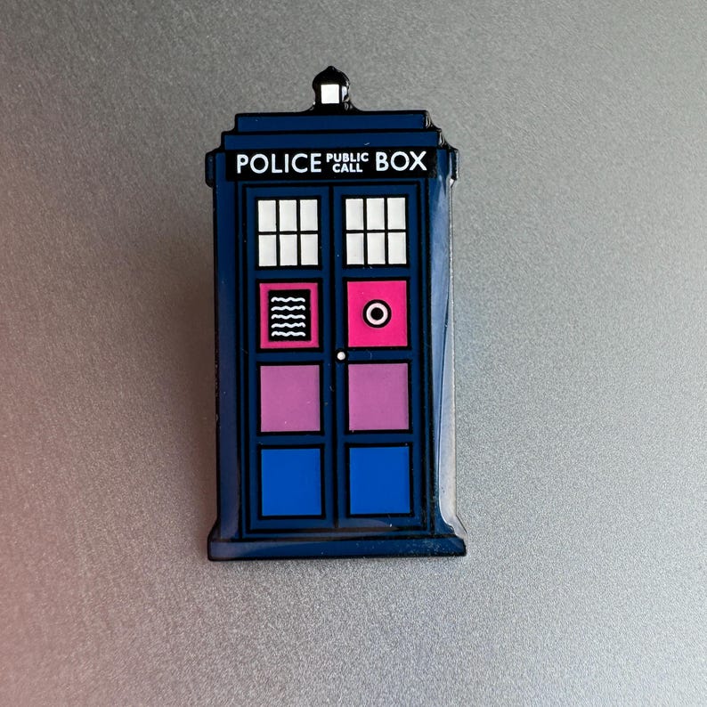 Pode incluir: Um &iacute;man azul da cabine telef&oacute;nica com o texto "POLICE PUBLIC CALL BOX". O &iacute;man apresenta um design retangular com janelas brancas, um c&iacute;rculo rosa e linhas onduladas. Os pain&eacute;is das portas s&atilde;o azuis e rosa.