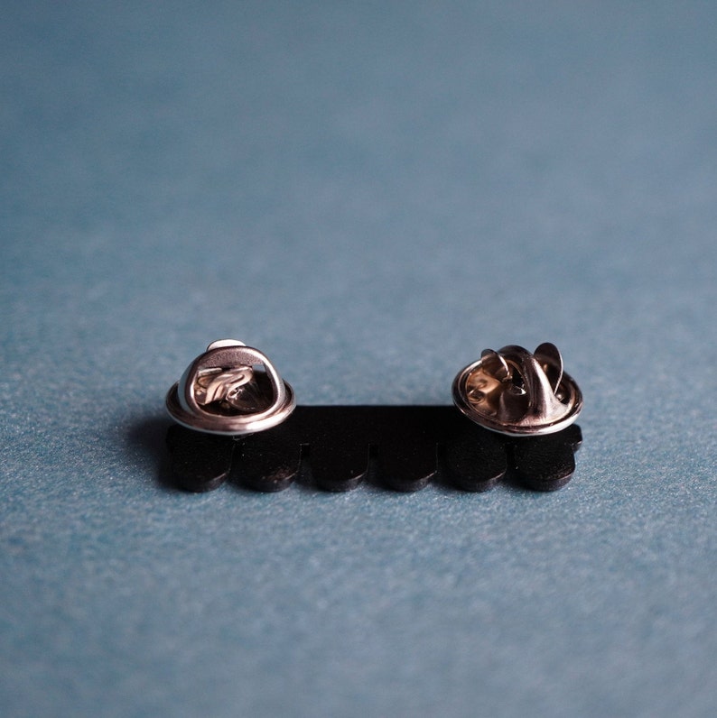 K&ouml;nnte beinhalten: R&uuml;ckansicht von zwei silbernen Metall-Stiftverschl&uuml;ssen, die an einer schwarzen Kunststoffunterlage mit gewellten Kanten befestigt sind.