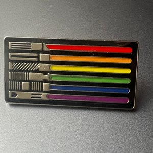 May include: A silver pin with a rainbow design. The pin features seven horizontal stripes in the colours of the rainbow, with a black background. The pin is shaped like a rectangle and has a silver finish.