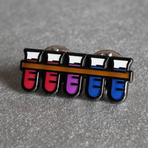 Pode incluir: Um alfinete de metal com cinco tubos de ensaio cheios de líquidos de diferentes cores. Os tubos de ensaio estão presos a uma barra de metal com uma faixa dourada. Os tubos de ensaio estão cheios de líquidos vermelho, rosa, roxo, azul e azul.