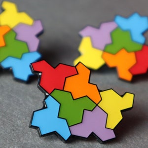 May include: Three colourful enamel pins in a jigsaw piece design. Each pin features a unique arrangement of vibrant colours including red, orange, yellow, green, blue, and purple, outlined in black. The pins are displayed on a grey surface.