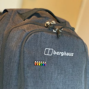 K&ouml;nnte beinhalten: Ein dunkelgrauer Rucksack mit dem Berghaus-Logo und einem Regenbogen-Patch. Der Rucksack hat eine Rei&szlig;verschlusstasche auf der Vorderseite.
