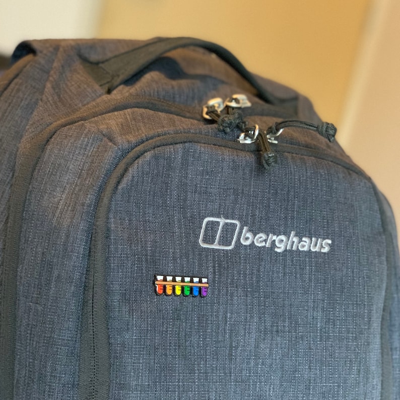 K&ouml;nnte beinhalten: Ein dunkelgrauer Rucksack mit dem Berghaus-Logo und einem Regenbogen-Patch. Der Rucksack hat eine Rei&szlig;verschlusstasche auf der Vorderseite.