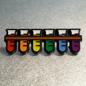 K&ouml;nnte beinhalten: Eine Regenbogen-Emaille-Anstecknadel mit sieben Reagenzgl&auml;sern in einem Gestell. Die R&ouml;hrchen sind mit roter, orangefarbener, gelber, gr&uuml;ner, blauer, indigofarbenen und violetter Fl&uuml;ssigkeit gef&uuml;llt.