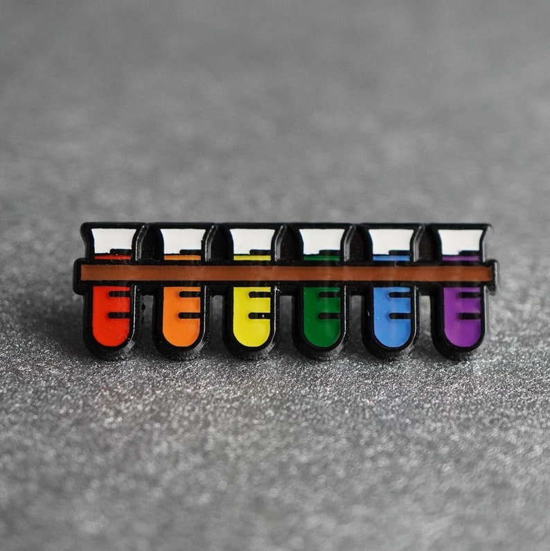 K&ouml;nnte beinhalten: Ein Satz von sieben Emaille-Pins in Form von Reagenzgl&auml;sern, jedes in einer anderen Farbe des Regenbogens. Die Pins sind in einer Reihe auf einer braunen Oberfl&auml;che angeordnet.