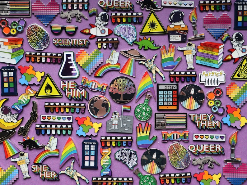 Pode incluir: Uma cole&ccedil;&atilde;o de alfinetes de esmalte coloridos com v&aacute;rios designs, incluindo arco-&iacute;ris, astronautas, c&eacute;rebros e textos como "SCIENTIST", "HE HIM", "THEY THEM" e "QUEER". Os alfinetes est&atilde;o dispostos sobre um fundo roxo.