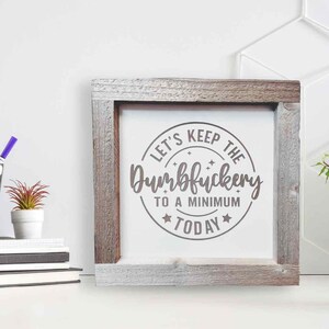 May include: A rustic wooden framed sign with a white background and a circle with the text "Let's Keep the Dumbfuckery to a Minimum Today" in brown lettering.