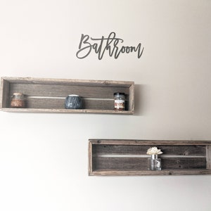 May include: Two rustic wooden shelves with a sign that reads "Bathroom" above them. The top shelf has a small glass jar, a black ceramic container, and a small glass jar with a label. The bottom shelf has a small glass bottle with a white flower inside.
