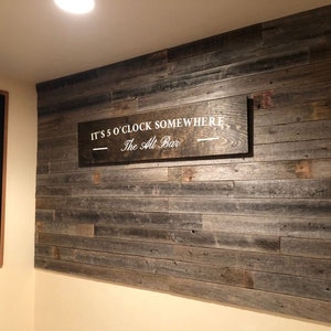 K&ouml;nnte beinhalten: Ein Holzschild mit dem Text "It's 5 o'clock somewhere - The Alt Bar" h&auml;ngt an einer Wand mit Holzverkleidung aus recyceltem Holz.