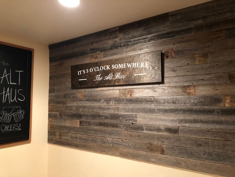 K&ouml;nnte beinhalten: Ein Holzschild mit dem Text "It's 5 o'clock somewhere - The Alt Bar" h&auml;ngt an einer Wand mit Holzverkleidung aus recyceltem Holz.