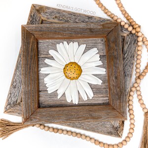 May include: A white daisy with a yellow center is mounted on a rustic wooden frame. The frame is double layered and has a distressed wood finish. The text "Kendra's Got Wood" is visible on the top of the frame.