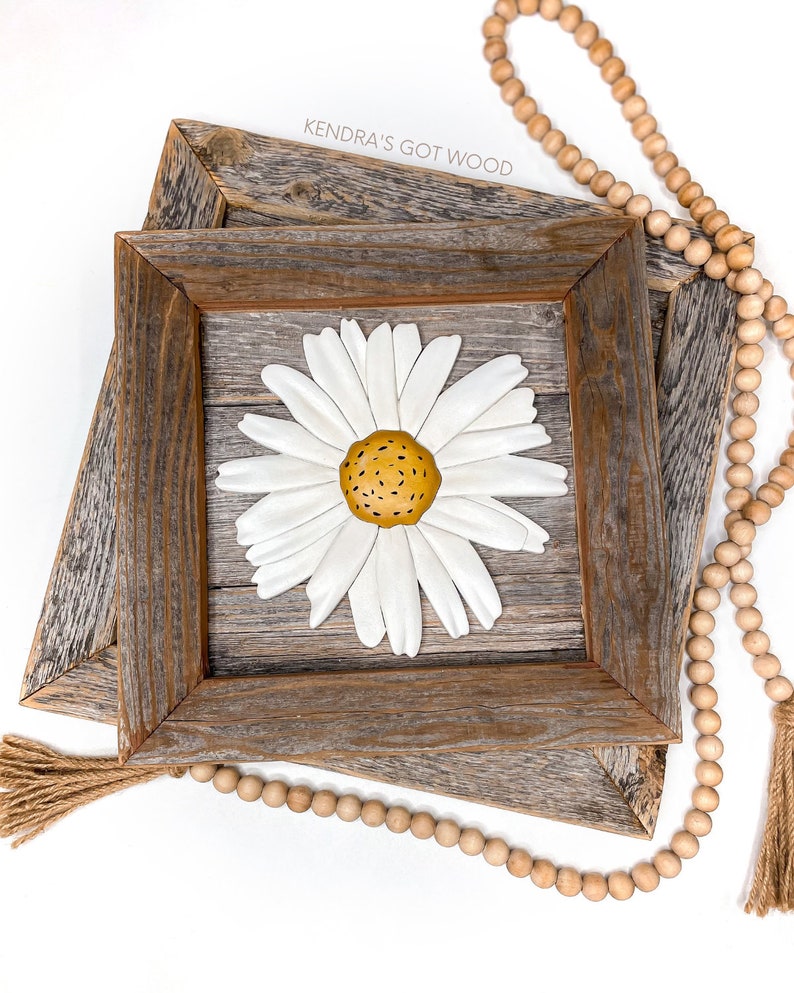 May include: A white daisy with a yellow center is mounted on a rustic wooden frame. The frame is double layered and has a distressed wood finish. The text "Kendra's Got Wood" is visible on the top of the frame.