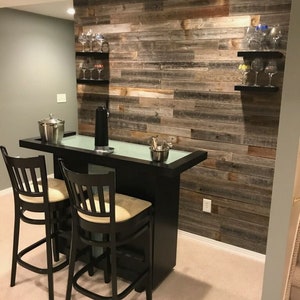 K&ouml;nnte beinhalten: Eine schwarze Bar mit zwei Barhockern und einer Glasplatte. Die Bar steht vor einer Wand mit wiederverwendeten Holzdielen. An der Wand befinden sich zwei Regale mit Weingl&auml;sern.